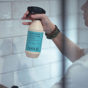 Hand holding a bottle of 'HOME' bathroom and shower cabin cleaner against a tiled bathroom background.