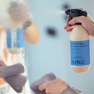 Person cleaning a mirror with a spray bottle labeled 'Lush Home' in a blurred indoor setting.